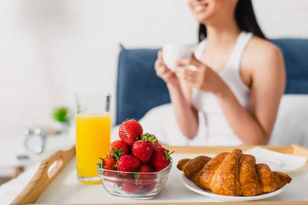 Selektiver Fokus von Erdbeeren, Orangensaft und Croissant auf Tablett in der Nähe einer Frau, die eine Tasse Kaffee auf dem Bett hält — Stockfoto