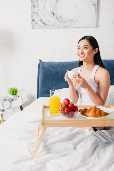 Foco seletivo de mulher asiática positiva segurando xícara de café perto do café da manhã na bandeja na cama — Fotografia de Stock