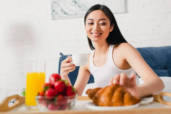 Избирательный фокус улыбающейся девушки, принимающей круассан во время завтрака в постели — стоковое фото