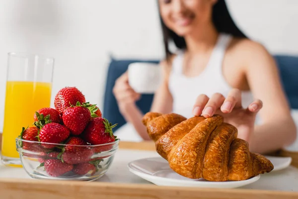 Selektiver Fokus der Frau, die Croissant in der Nähe von Erdbeeren und ein Glas Orangensaft auf dem Frühstückstablett nimmt — Stockfoto