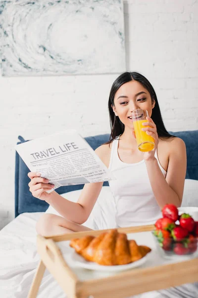 Selektiver Fokus einer lächelnden asiatischen Frau, die Zeitung mit Reisebuchstaben liest und ein Glas Orangensaft auf dem Bett hält — Stockfoto