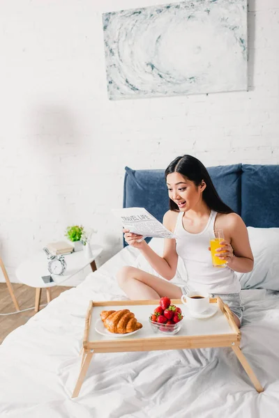 Alegre ásia menina leitura jornal perto pequeno-almoço no pequeno-almoço bandeja no cama no casa — Fotografia de Stock