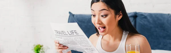 Panorâmica tiro de animado asiático mulher ler jornal com viagem vida letras e segurando vidro de suco de laranja — Fotografia de Stock