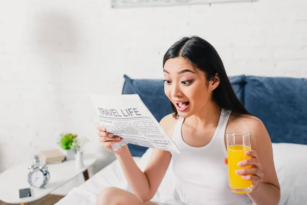 Excited asian woman reading newspaper with travel life lettering and holding glass of orange juice on bed — Stock Photo
