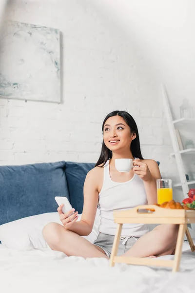 Enfoque selectivo de sonriente mujer asiática sosteniendo taza de café y teléfono inteligente cerca del desayuno en la cama - foto de stock