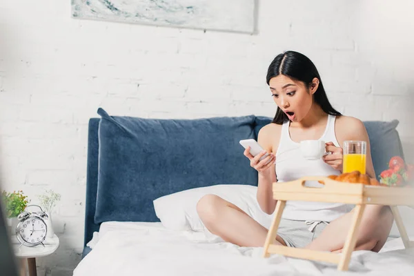 Enfoque selectivo de mujer asiática conmocionada usando teléfono inteligente durante el desayuno en el dormitorio - foto de stock