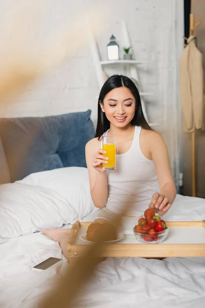 Selektiver Fokus einer lächelnden asiatischen Frau mit einem Glas Orangensaft und Erdbeere im Bett — Stockfoto