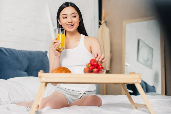 Избранное внимание веселой азиатской девушки, держащей стакан апельсинового сока рядом с завтраком с клубникой на кровати — стоковое фото