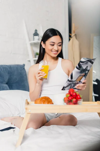 Selective focus of smiling asian woman reading magazine during breakfast with orange juice on bed — Stock Photo