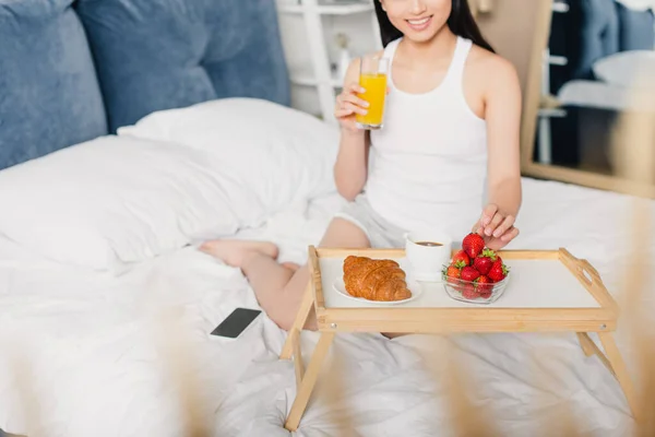 Vista recortada de una mujer sonriente sosteniendo un vaso de jugo de naranja y tomando fresa cerca de croissant y café en la bandeja del desayuno en la cama - foto de stock