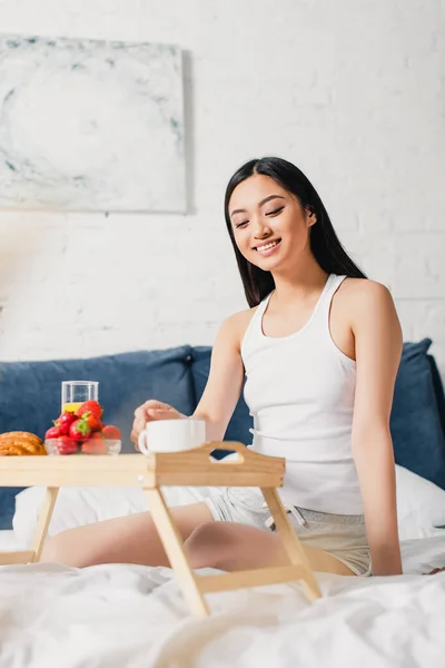 Selective focus of smiling asian woman sitting near breakfast tray on bed at morning — Stock Photo