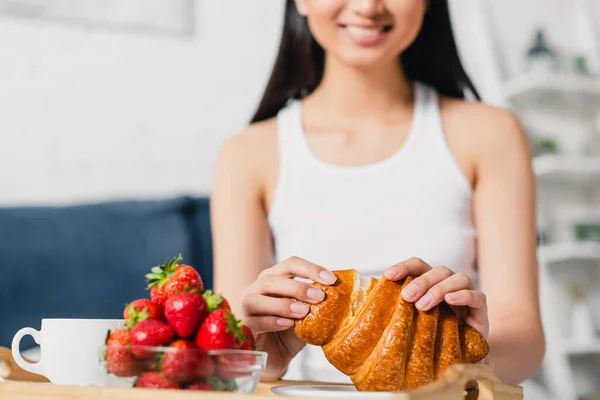 Selective focus of smiling woman holding croissant near strawberries and coffee in bedroom — Stock Photo