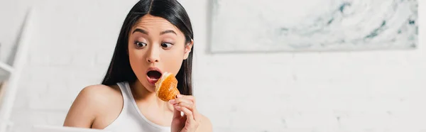 Panoramic shot of shocked asian woman holding piece of croissant at home — Stock Photo