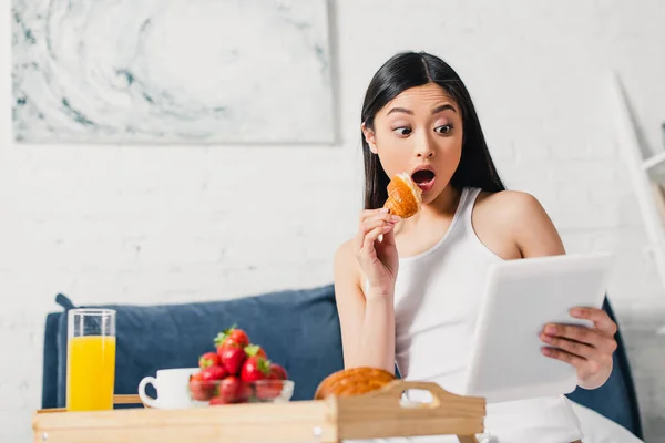 Foco seletivo de mulher asiática surpresa segurando pedaço de croissant e tablet digital perto do café da manhã na cama — Fotografia de Stock