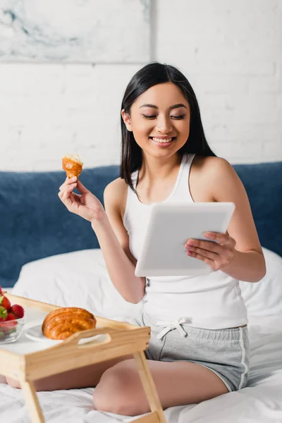Focus selettivo di sorridere donna asiatica utilizzando tablet digitale vicino a colazione sul letto — Foto stock