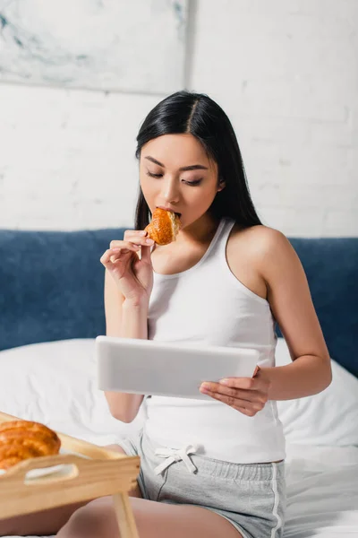 Selective focus of attractive asian girl eating croissant and using digital tablet on bed — Stock Photo