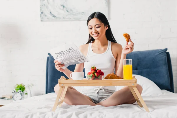 Foco seletivo de mulher asiática alegre ler jornal e segurando pedaço de croissant na cama — Fotografia de Stock