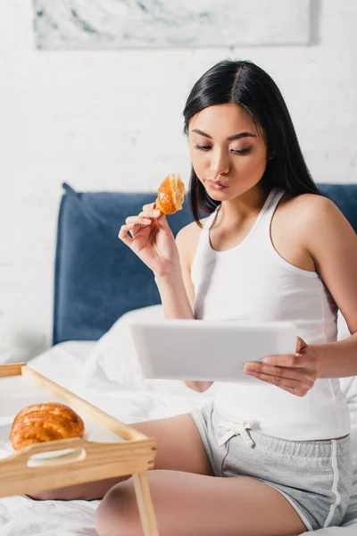 Foco seletivo de mulher asiática usando tablet digital e comer croissant perto bandeja de café da manhã na cama — Fotografia de Stock