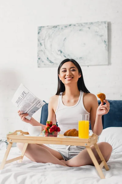 Selektiver Fokus eines asiatischen Mädchens, das in die Kamera lächelt, während es Zeitung und Croissant auf dem Bett hält — Stockfoto