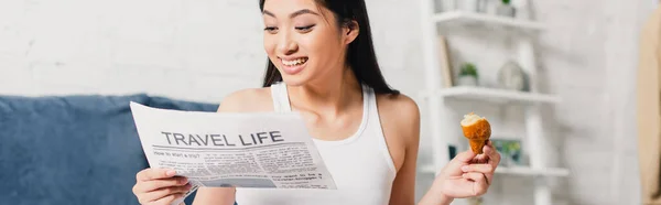 Panoramic crop of smiling asian woman holding piece of croissant while reading newspaper at home — Stock Photo