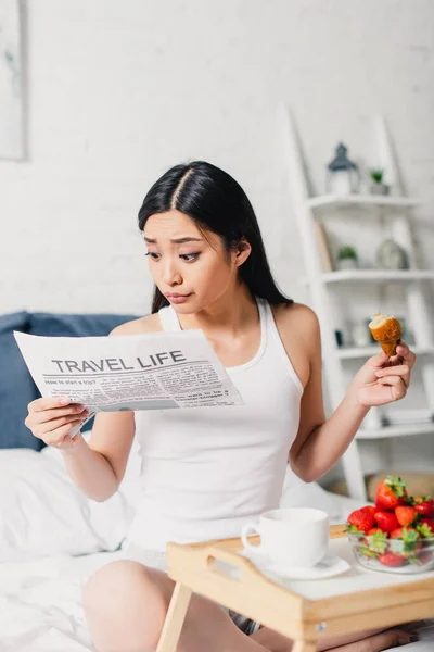 Selective focus of asian woman reading news and holding croissant near strawberries and coffee on breakfast tray on bed — Stock Photo