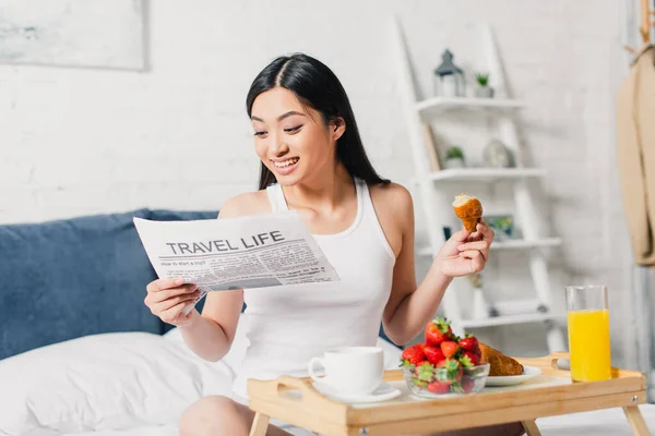 Enfoque selectivo de alegre chica asiática leyendo noticias cerca del desayuno con fresas frescas y croissant en la cama - foto de stock