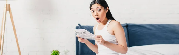 Horizontal cosecha de shocked asiático mujer holding periódico en cama - foto de stock