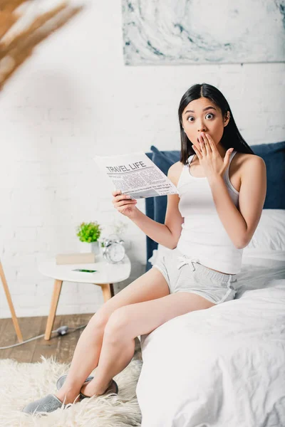 Selektiver Fokus einer schockierten asiatischen Frau, die Mund mit der Hand bedeckt, während sie Zeitung auf dem Bett hält — Stockfoto