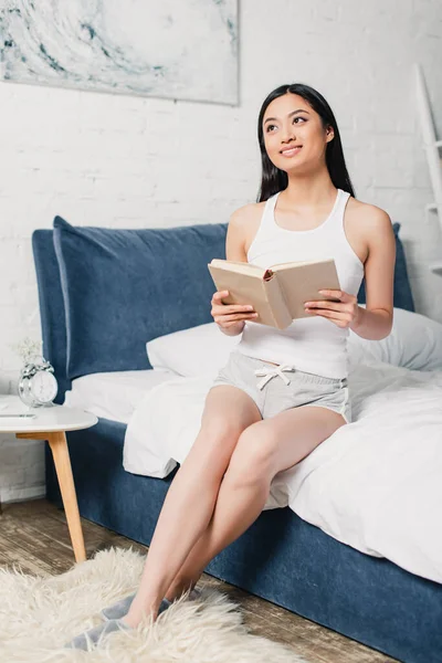 Positive asiatische Frau hält offenes Buch auf dem Bett — Stockfoto