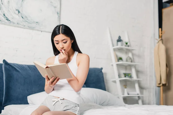 Концентрована азіатська жінка читає книгу, сидячи на ліжку — стокове фото
