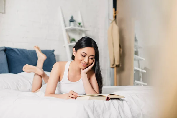 Выборочный фокус улыбающейся азиатской девушки, читающей книгу, лежащей дома на кровати — стоковое фото