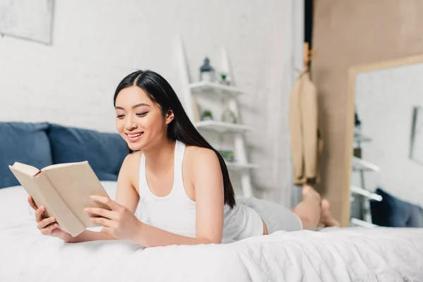 Atractiva mujer asiática sonriendo mientras lee libro en la cama - foto de stock