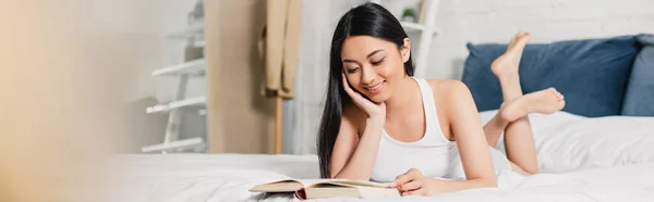 Horizontal image of smiling asian girl with hand near cheek reading book on bed — Stock Photo