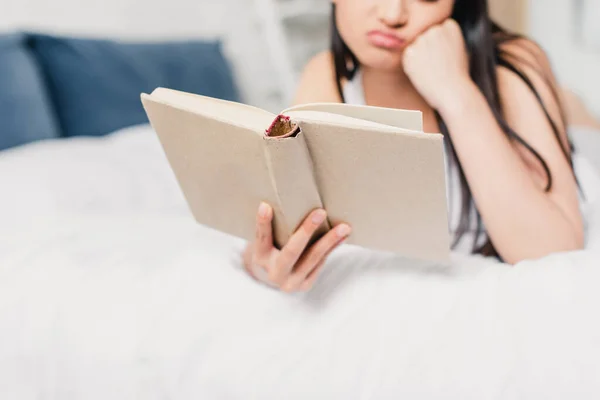 Vista ritagliata di donna con mano vicino guancia libro di lettura mentre sdraiato sul letto — Foto stock