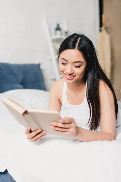 Enfoque selectivo de sonriente mujer asiática lectura libro en dormitorio - foto de stock