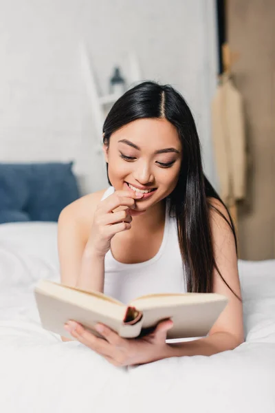 Selective focus of smiling asian woman with finger near lips reading book on bed — Stock Photo
