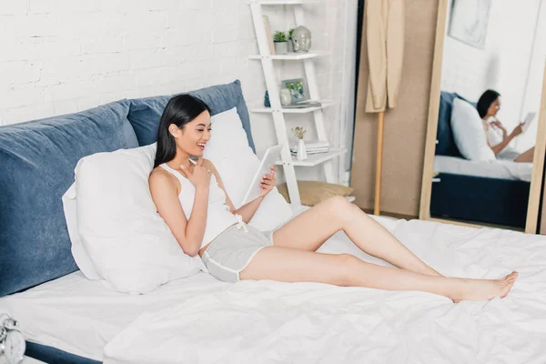 Foyer sélectif de gai asiatique fille en pyjama en utilisant tablette numérique sur lit — Photo de stock