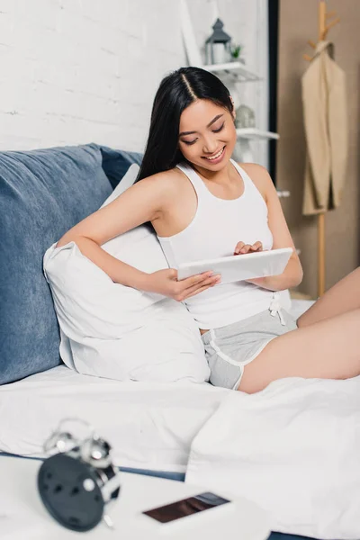 Selective focus of smiling asian girl using digital tablet while sitting on bed — Stock Photo