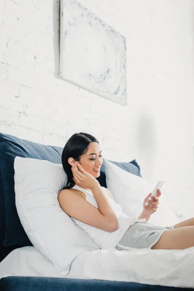 Hermosa mujer asiática usando teléfono inteligente y sonriendo en la cama - foto de stock