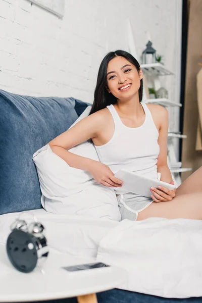 Selective focus of asian girl smiling at camera while holding digital tablet on bed — Stock Photo