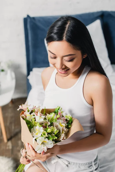 Smiling asian woman holding bouquet while sitting on bed — Stock Photo