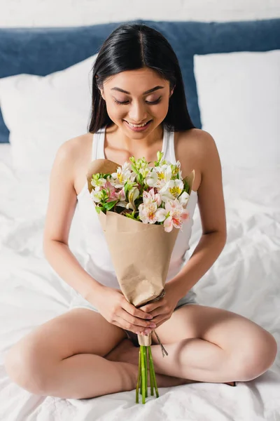 Hermosa chica asiática sonriendo mientras sostiene ramo en la cama - foto de stock
