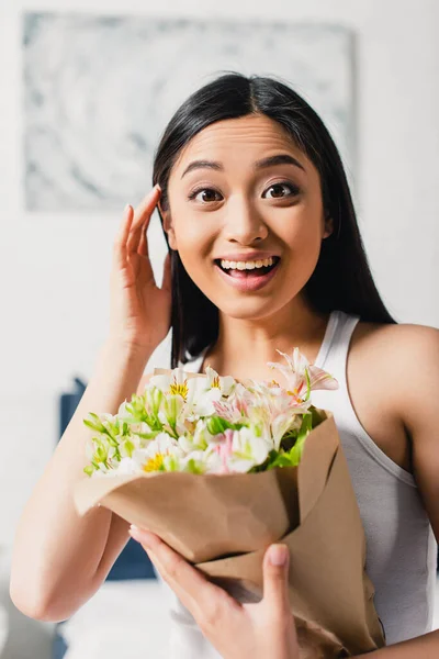 Selective focus of cheerful asian girl holding bouquet and looking at camera at home — Stock Photo