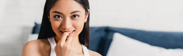 Horizontal colheita de mulher asiática com o dedo perto de lábios sorrindo para a câmera no quarto — Fotografia de Stock