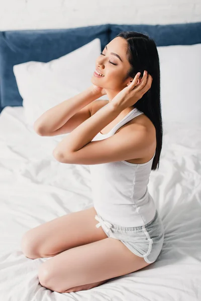 Smiling asian girl touching hair while sitting on bed — Stock Photo