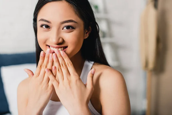 Cheerful asian girl with hands near mouth smiling at camera at home — Stock Photo