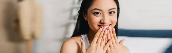 Colpo panoramico di donna asiatica positiva con le mani vicino alla bocca guardando la fotocamera a casa — Foto stock