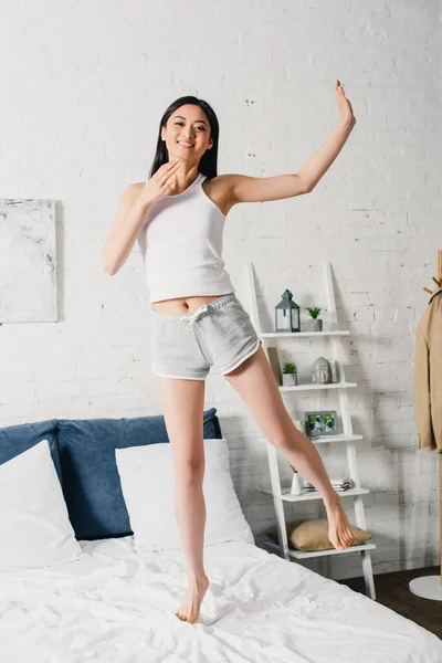 Positive asian woman smiling at camera while standing on bed — Stock Photo