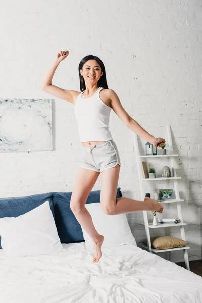 Cheerful asian woman in pajamas jumping on bed — Stock Photo