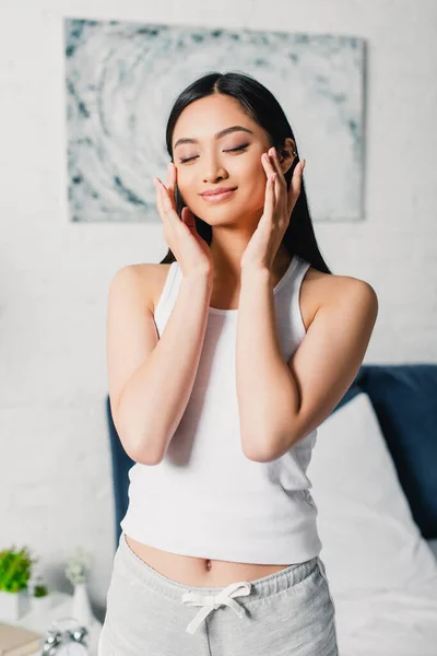 Young asian woman with closed eyes touching face in bedroom — Stock Photo
